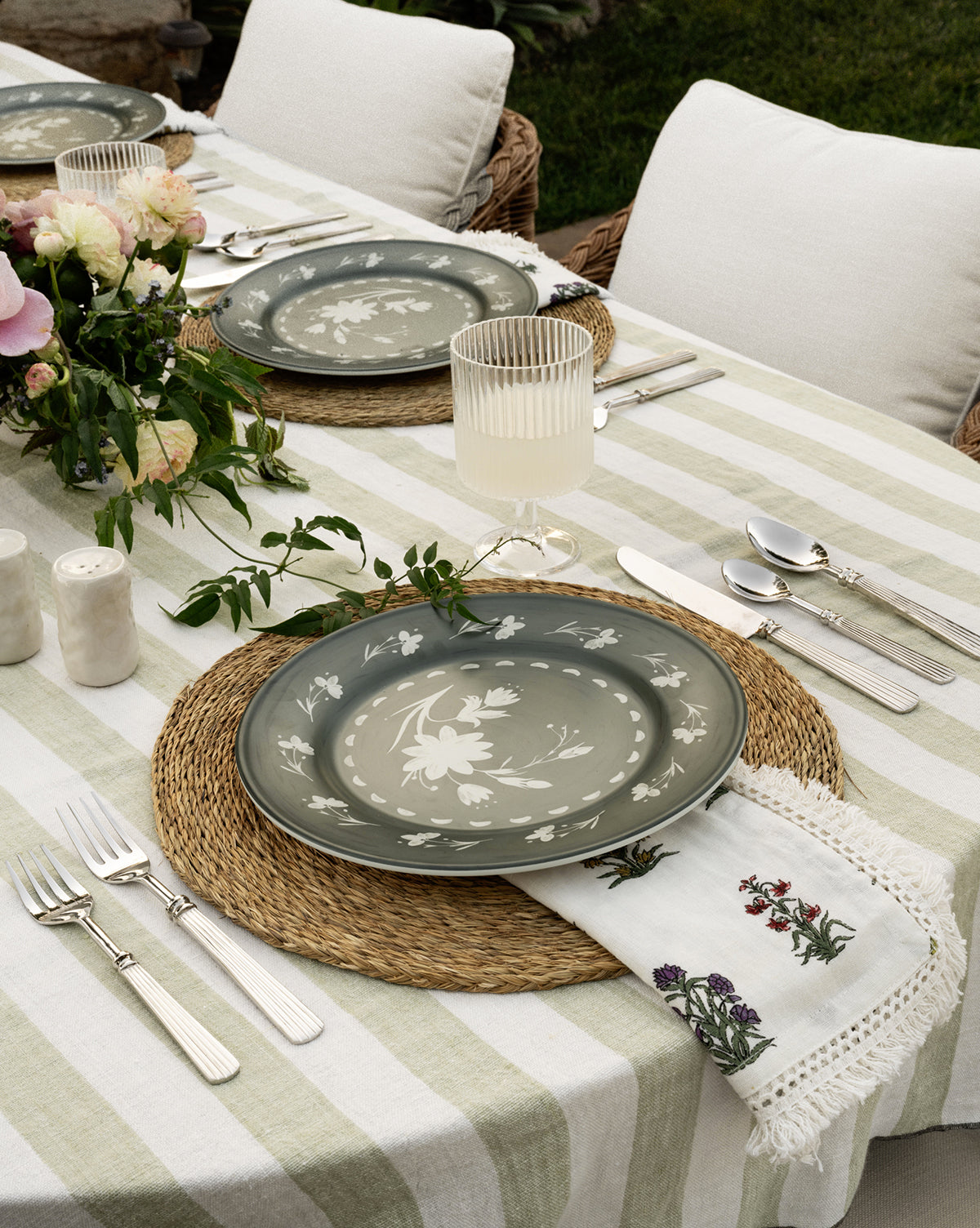The outdoor table set, featuring striped tablecloth, woven placemats, floral plates, embroidered napkins, silver cutlery, a floral centerpiece, and Sanibel Acrylic Goblets (Set of 4), creates an elegant and inviting setting.