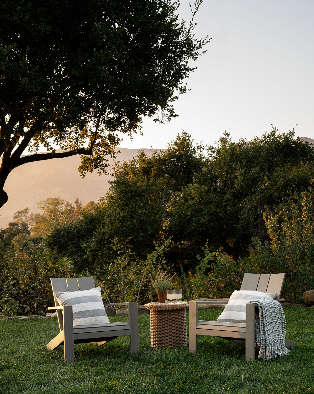 Two Triby Gray Teak Outdoor Lounge Chairs with cushions and throws sit on grass facing a small round wicker table with drinks and a plant, surrounded by greenery and trees, with mountains visible in the sunset—a relaxing outdoor setup, by McGee & Co.