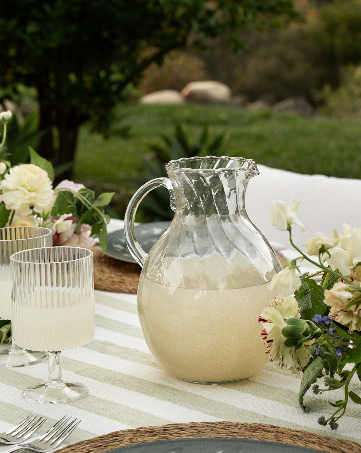 The Adderley Pitcher, made of mouth-blown glass and filled with a light beverage, sits on an outdoor table alongside two matching glasses, floral arrangements, and greenery in the background, by Vendixs