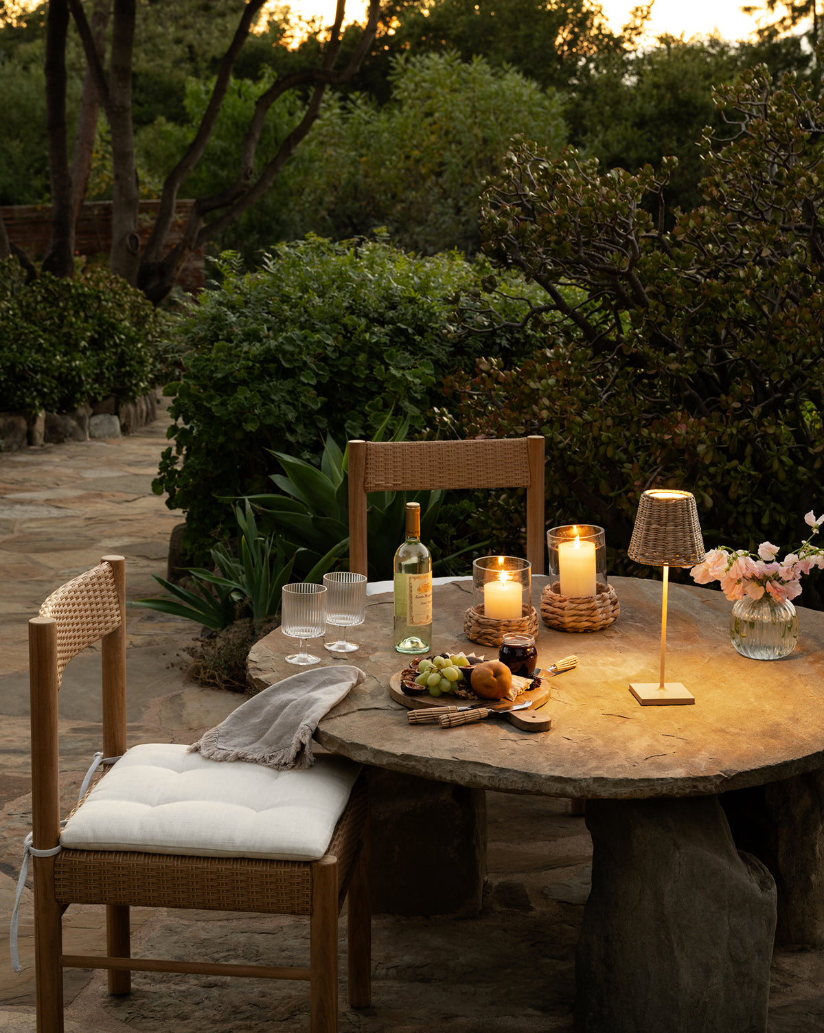 A rustic stone table outdoors, set for two with wine and cheese among greenery at dusk, features the Poldina Indoor/Outdoor Table Lamp, adding a modern touch to the inviting scene, by Vendixs
