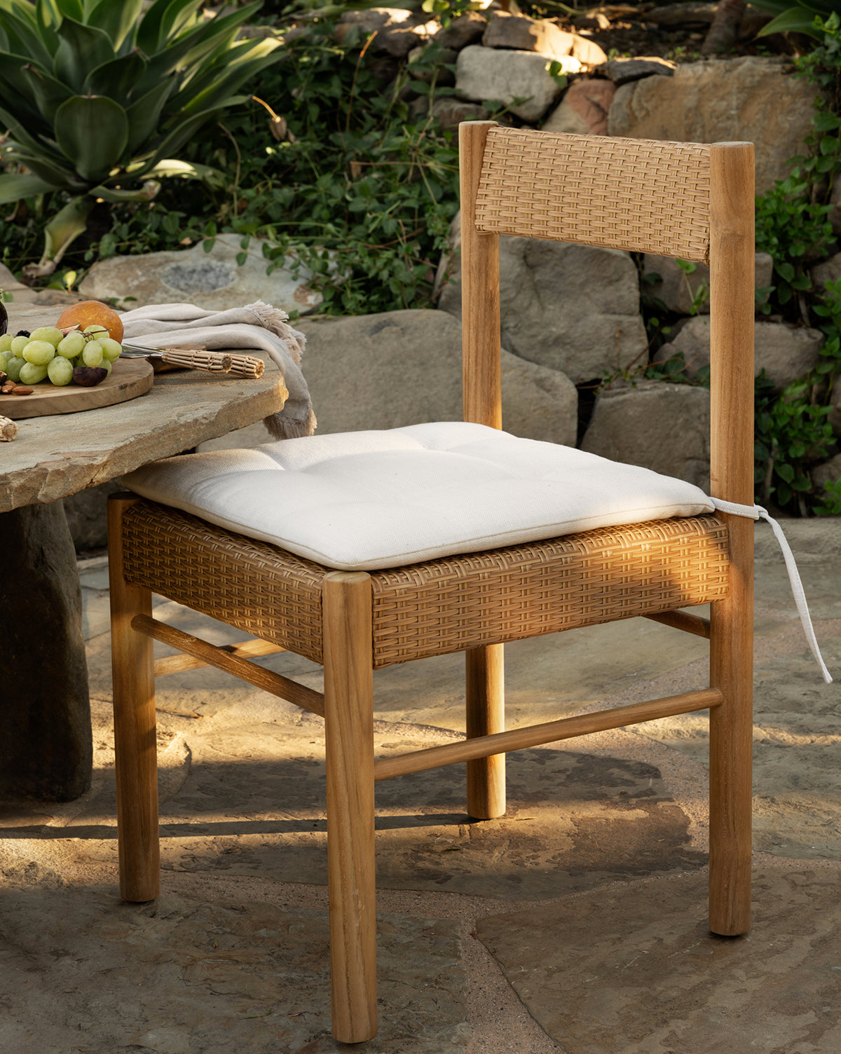 The Simeon Outdoor Dining Chair, featuring a woven rattan seat and backrest with a white cushion, sits by a stone table holding grapes in a garden surrounded by rocks and lush green plants, by Vendixs