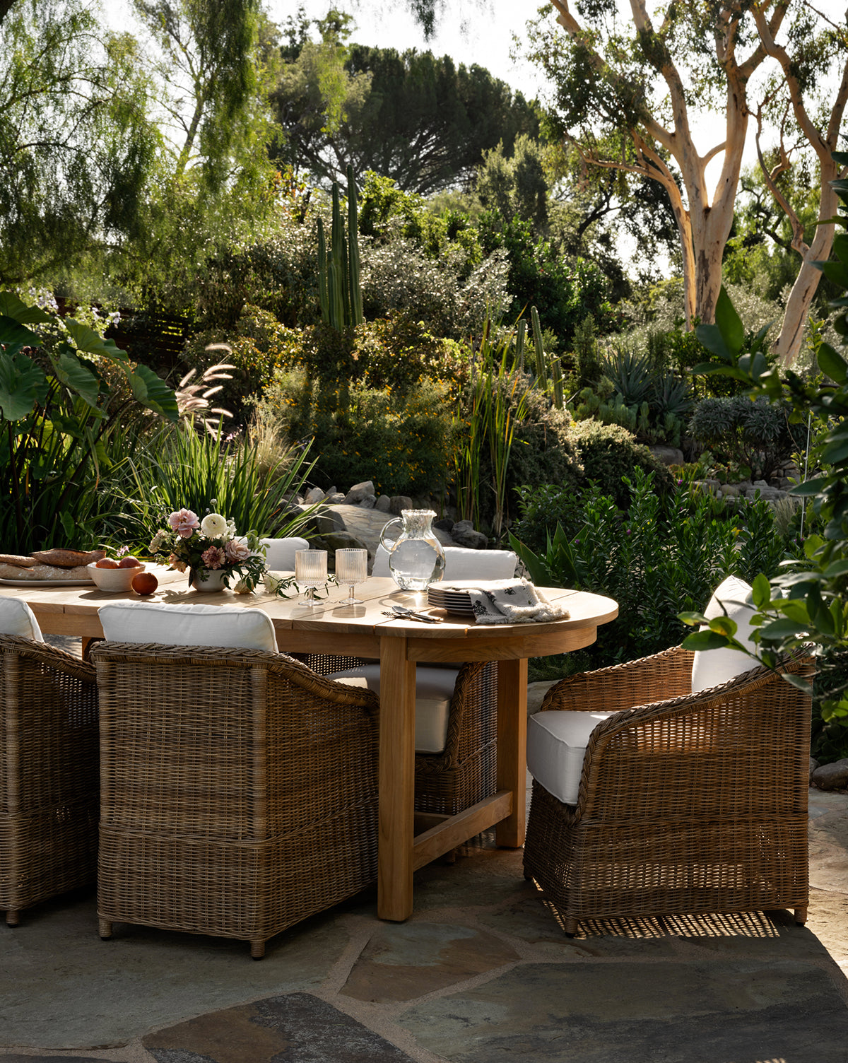 A round wooden table is set for a meal with Haviland Outdoor Dining Chairs featuring weather-resistant wicker and white cushions. The table has glassware, plates, napkins, and a floral centerpiece, surrounded by lush green patio plants by McGee & Co.