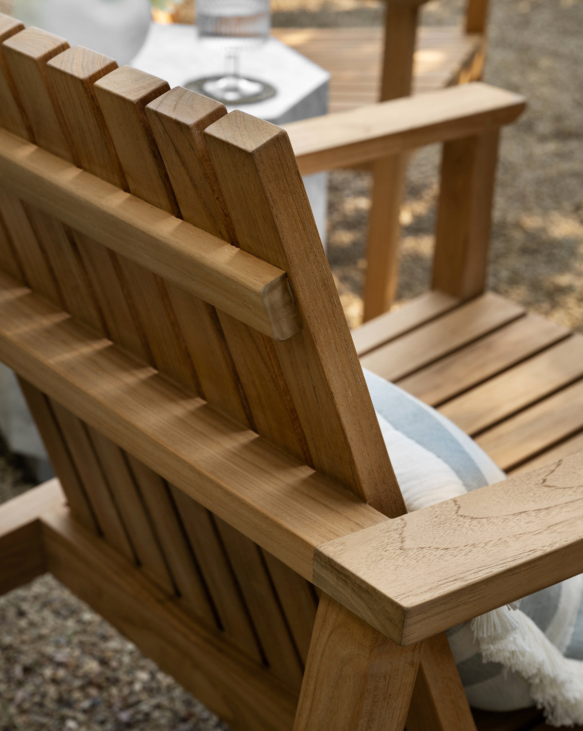 Close-up of the Gilda Outdoor Lounge Chair in teak wood, featuring a slatted back and seat with a blue and white striped cushion. In the background, a small table holds a glass and carafe on a gravel surface, by McGee & Co.