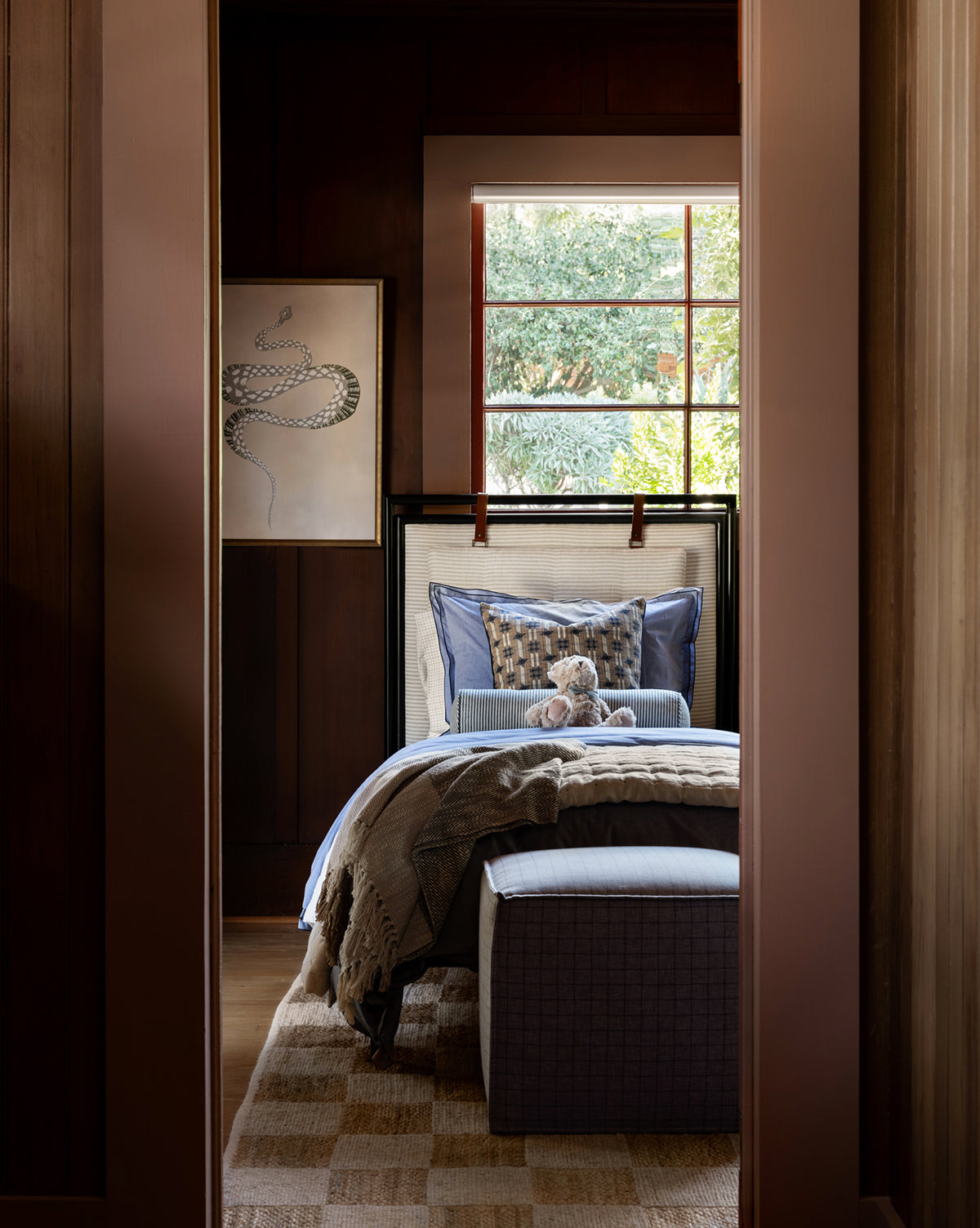 A cozy bedroom with wood-paneled walls, blue and beige bedding, and a teddy bear. A cushioned seat is nearby, while the Johannes Cube framed snake artwork hangs above. Natural light streams in from a window overlooking greenery by McGee & Co.