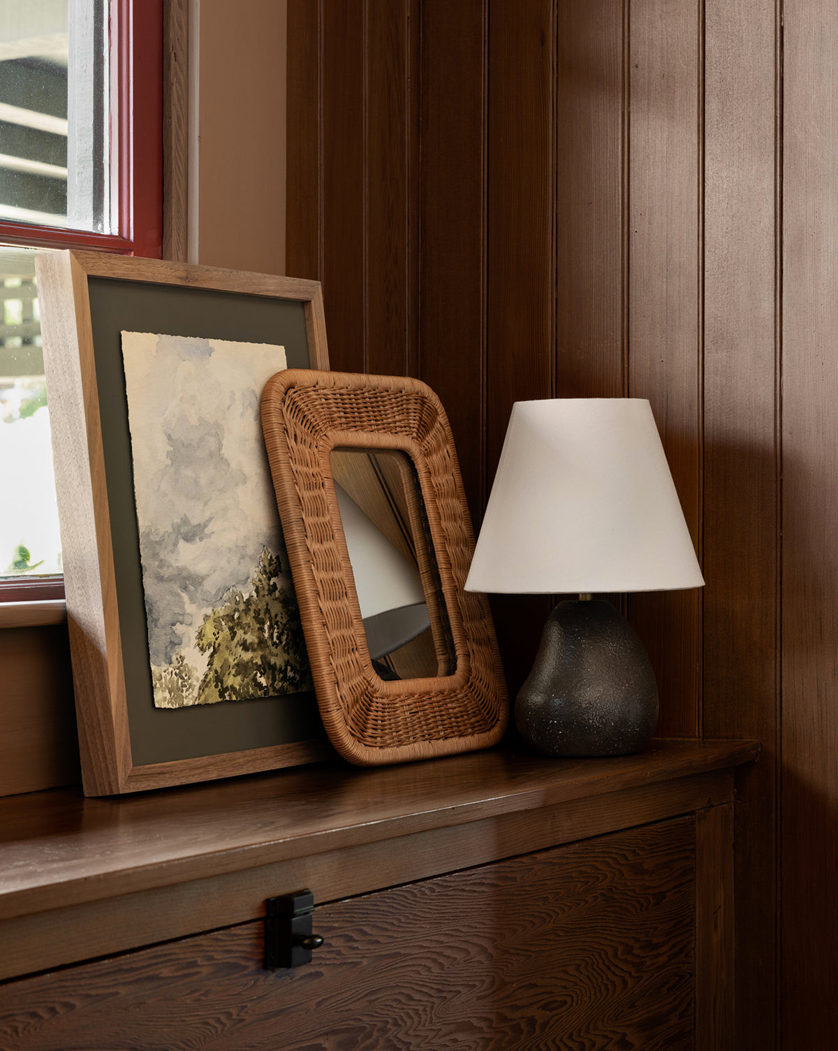 A framed painting, the Jera Shelf Mirror with a wicker frame, and a small table lamp with a white shade sit on a wooden surface against a paneled wall beside a window by Vendixs
