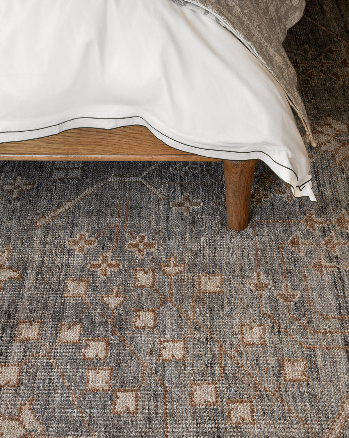 A corner of a bed with white and beige bedding sits on a wooden frame above the Elison Hand-Knotted Wool Rug, featuring geometric and floral motifs that highlight artisan craftsmanship, by Vendixs