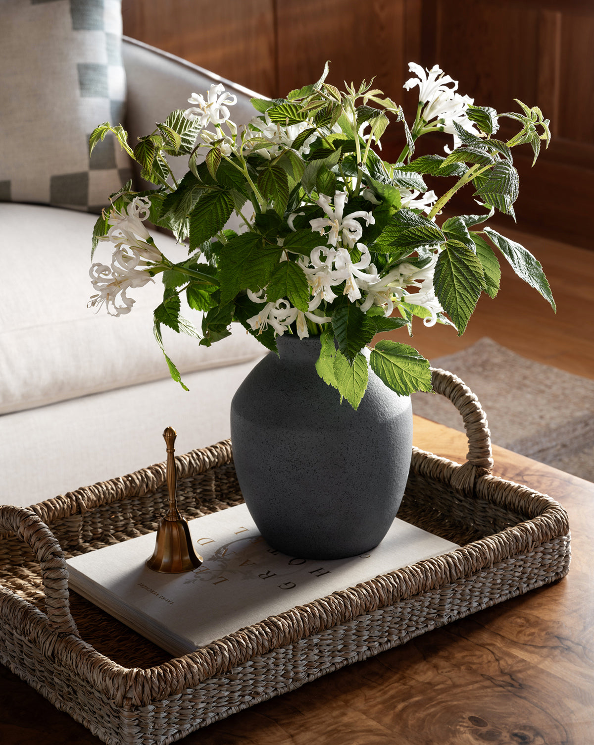 A woven tray on a wooden table holds the Delgado Vase with white flowers and green leaves, a small gold bell, and a closed book. In the background, there’s a beige sofa with a patterned pillow, by Vendixs