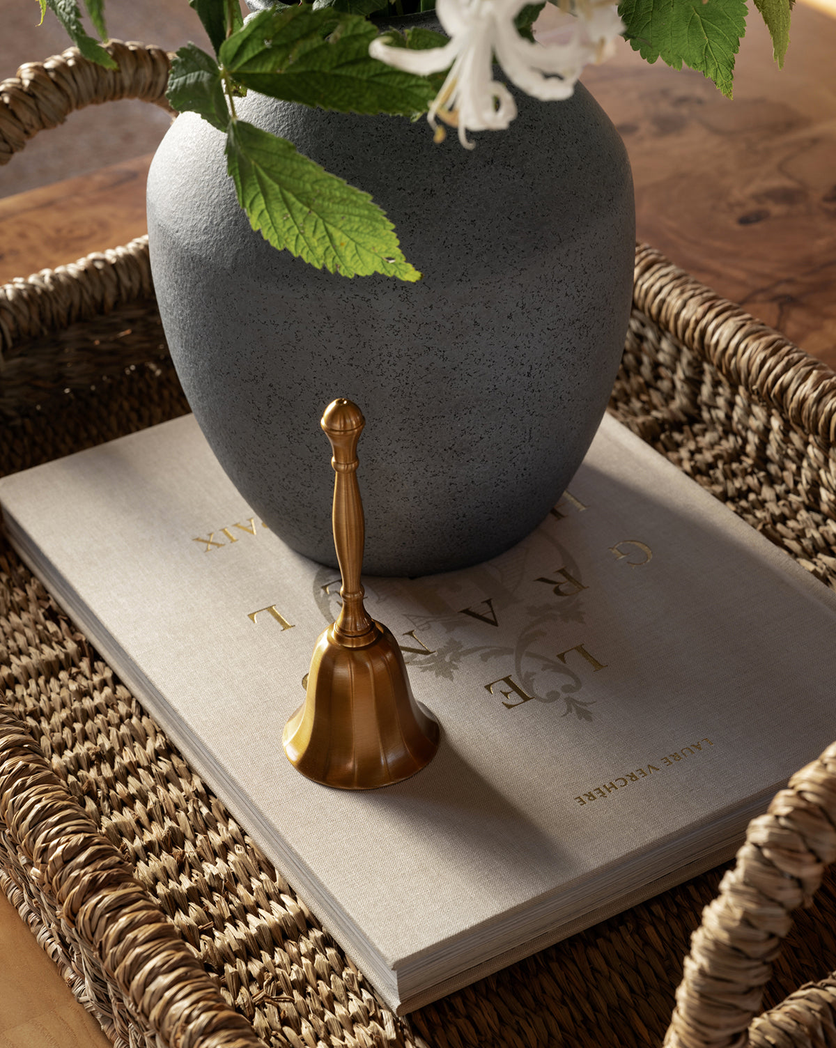 The Elsher Bell, crafted in golden antique brass, sits atop a large gray book with gold lettering beside a gray vase of white flowers and green leaves, all arranged in a woven tray for classic vintage decor, by Vendixs