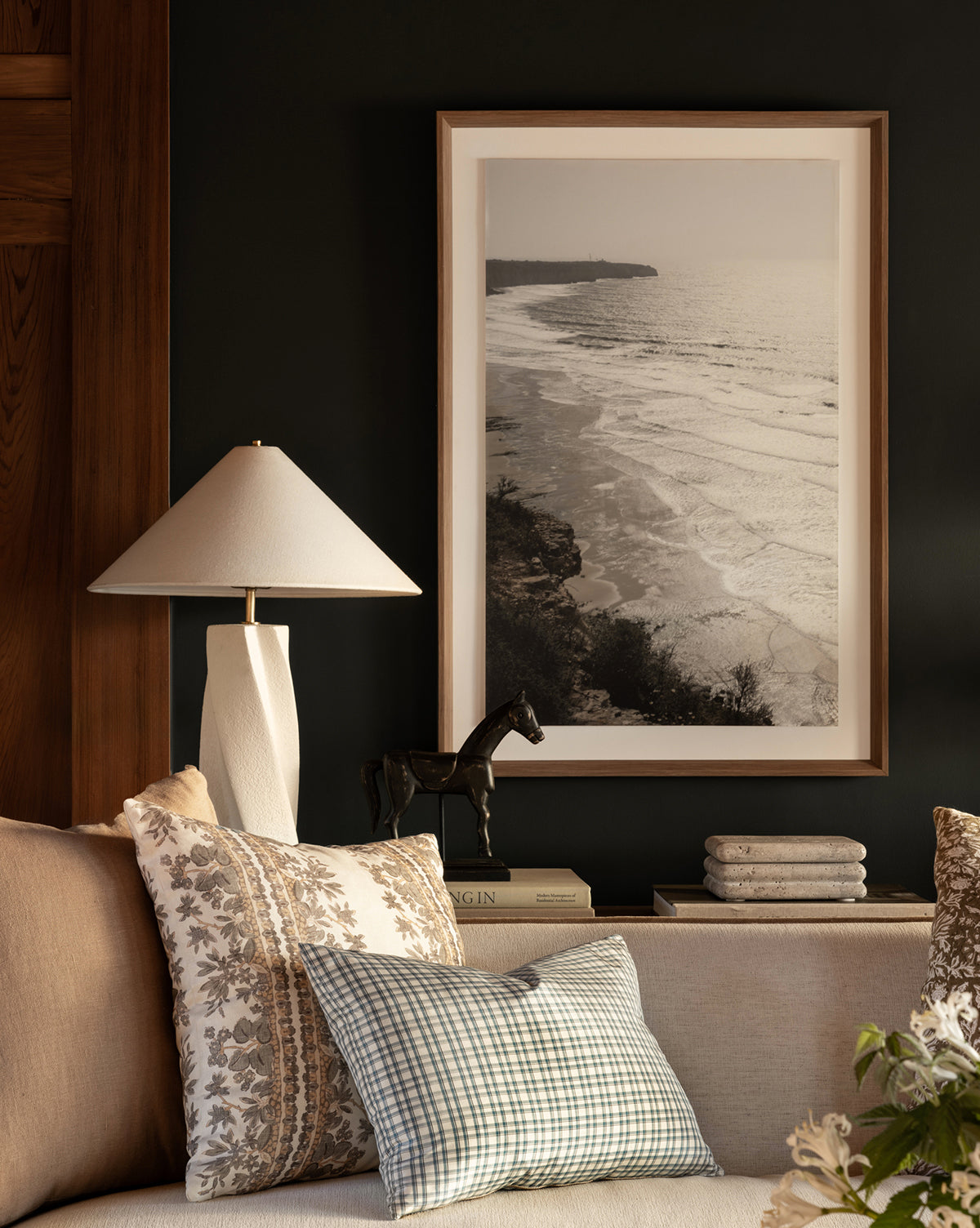 A cozy living room corner features a beige sofa, patterned pillows, stacked books, a horse figurine, and the Bettencourt Table Lamp with a linen shade next to a framed black-and-white coastal photo on a dark wall, by Vendixs