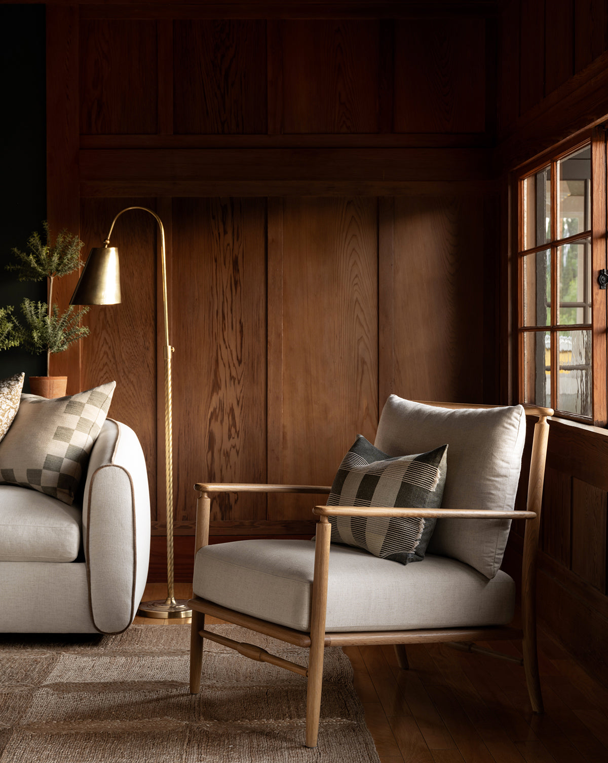 A cozy living room with wood-paneled walls, a Fenwick Lounge Chair, matching sofa, patterned pillows, brass floor lamp, and sunlight pouring through a window onto the rug, by Vendixs