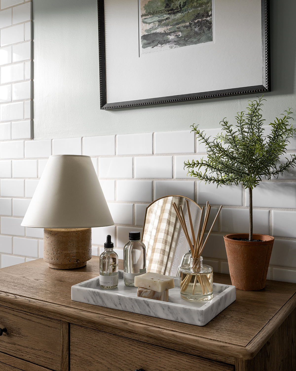 A wooden dresser topped with the Bronte Table Lamp, a potted plant, toiletries on a tray, and decorative accents sits against white subway tiles beneath a framed landscape painting by Vendixs