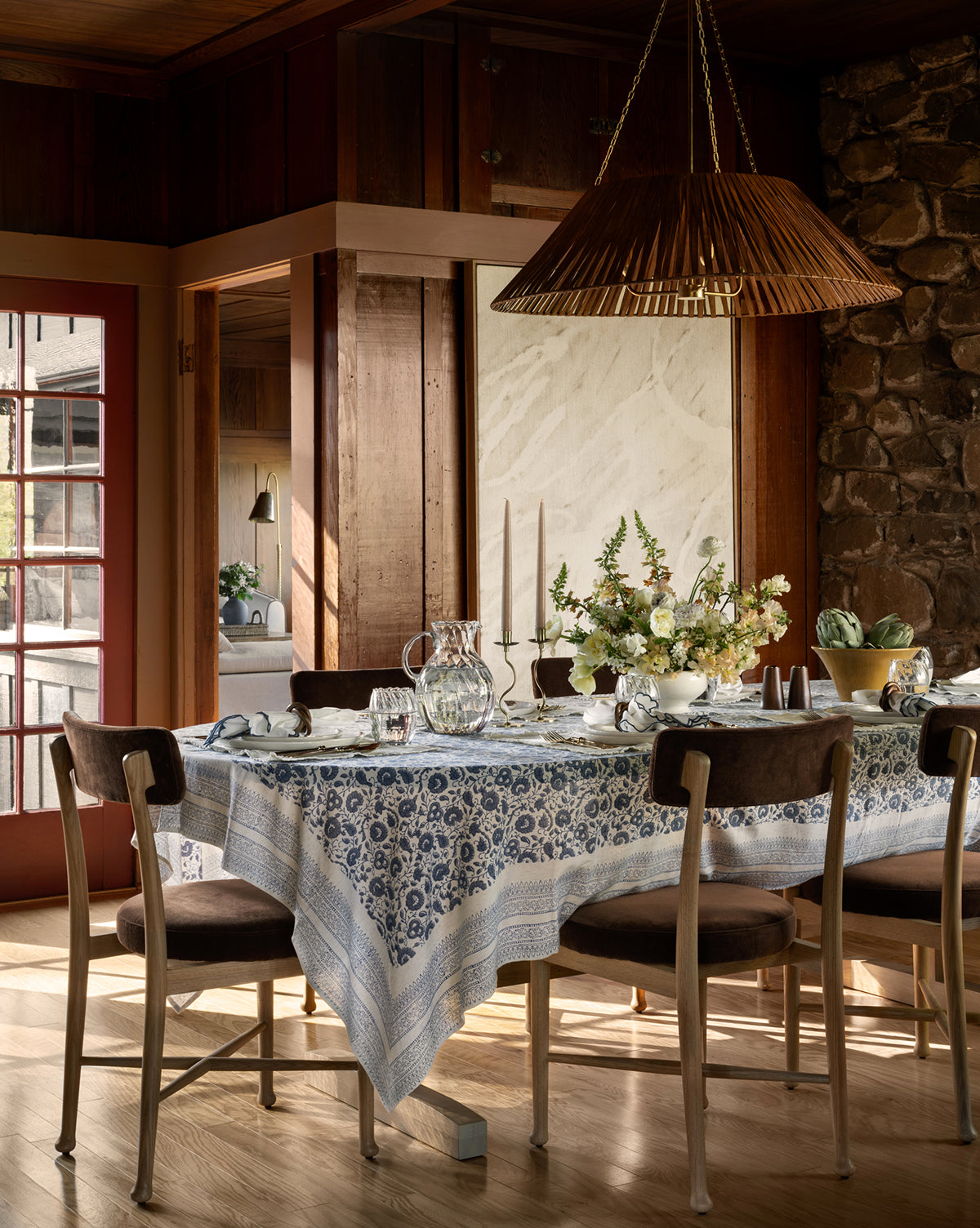 A rustic dining room features a wooden table draped with the Jennie Tablecloth, surrounded by wooden chairs, floral centerpieces, candles, glassware, and natural light streaming through large windows, by Vendixs