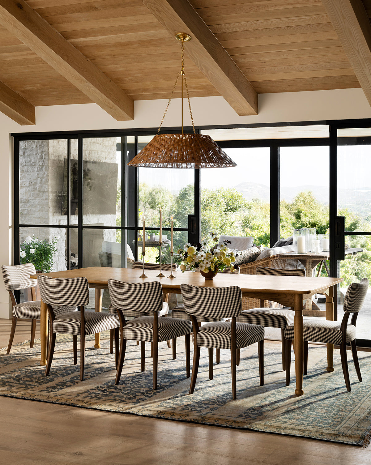 A bright dining room with a wooden table, eight Etienne Dining Chairs, a woven pendant light, floral centerpiece, large windows with greenery views, all set on a patterned rug beneath an exposed wood beam ceiling by McGee & Co.