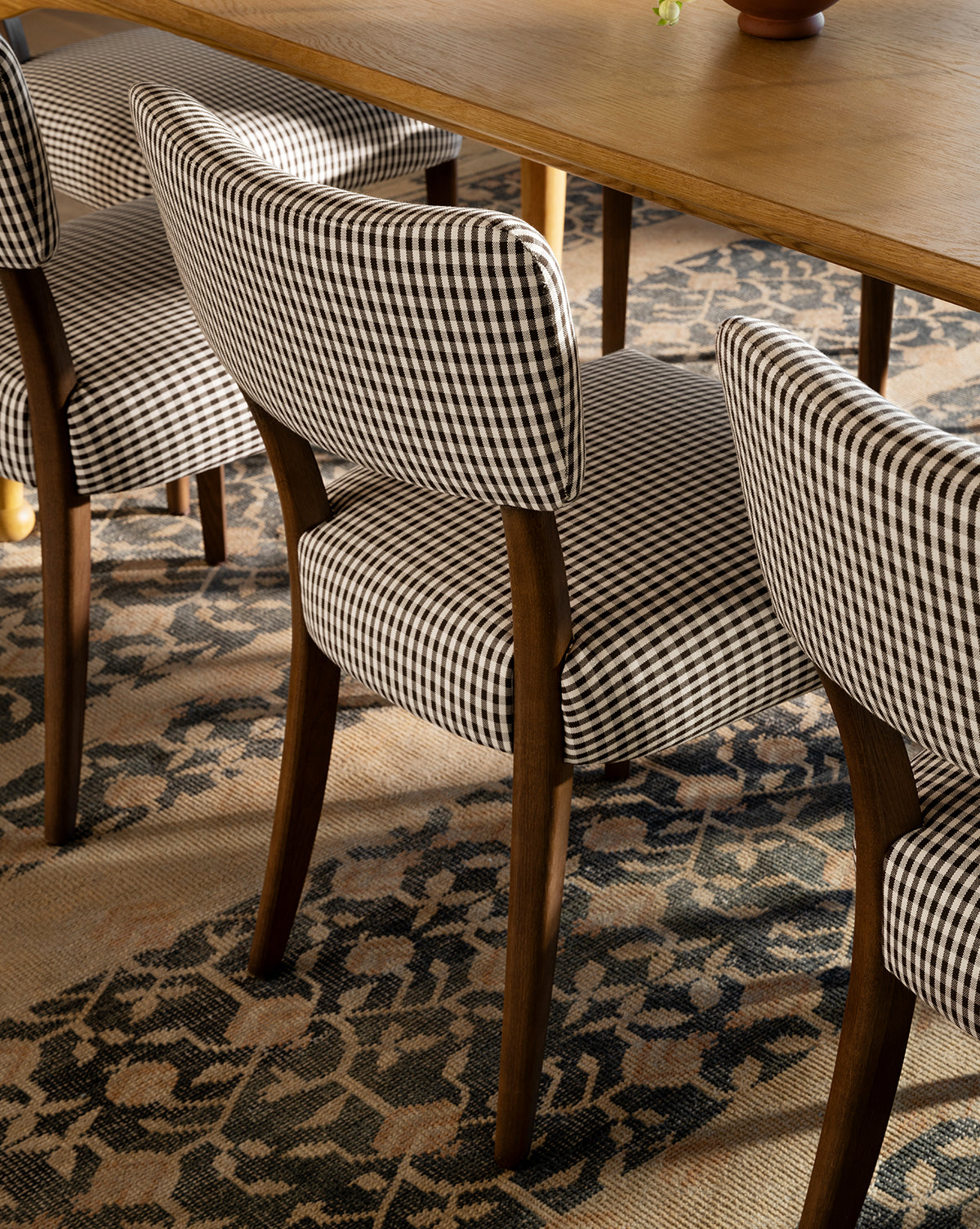 A close-up of Makerspalm Etienne Dining Chairs, featuring wooden legs and black-and-white checkered upholstery, arranged by a wooden dining table on a patterned carpet. Vendixs