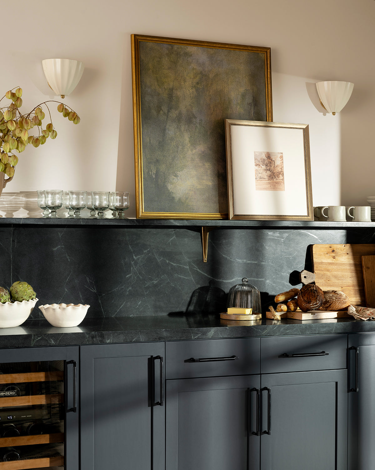 A modern kitchen with dark gray cabinets, black marble countertops, cutting boards, glassware, bowls, and the Harewood Park print in a distressed silver wood frame leaning on a shelf under soft wall sconces, by Vendixs