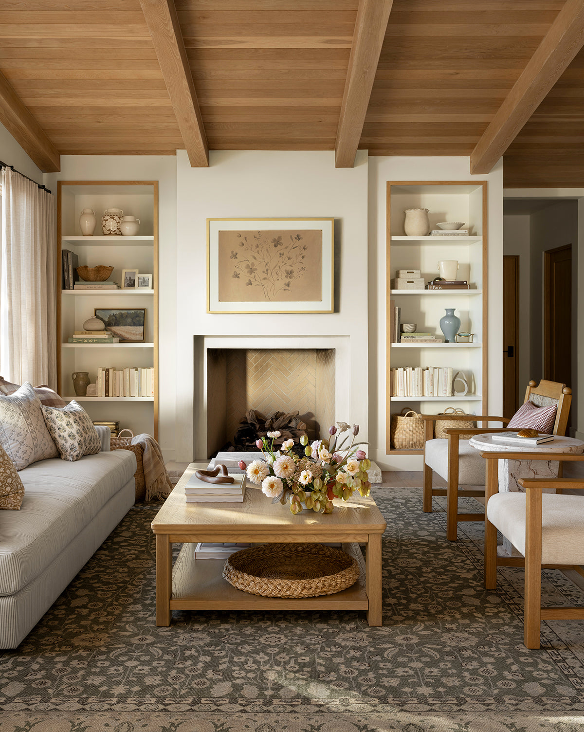 A cozy living room with wooden ceiling beams, built-in shelves, a framed artwork above the fireplace, a beige sofa, two armchairs, a wooden coffee table with flowers, and the Fernwood Hand-Knotted Wool Rug, by Vendixs