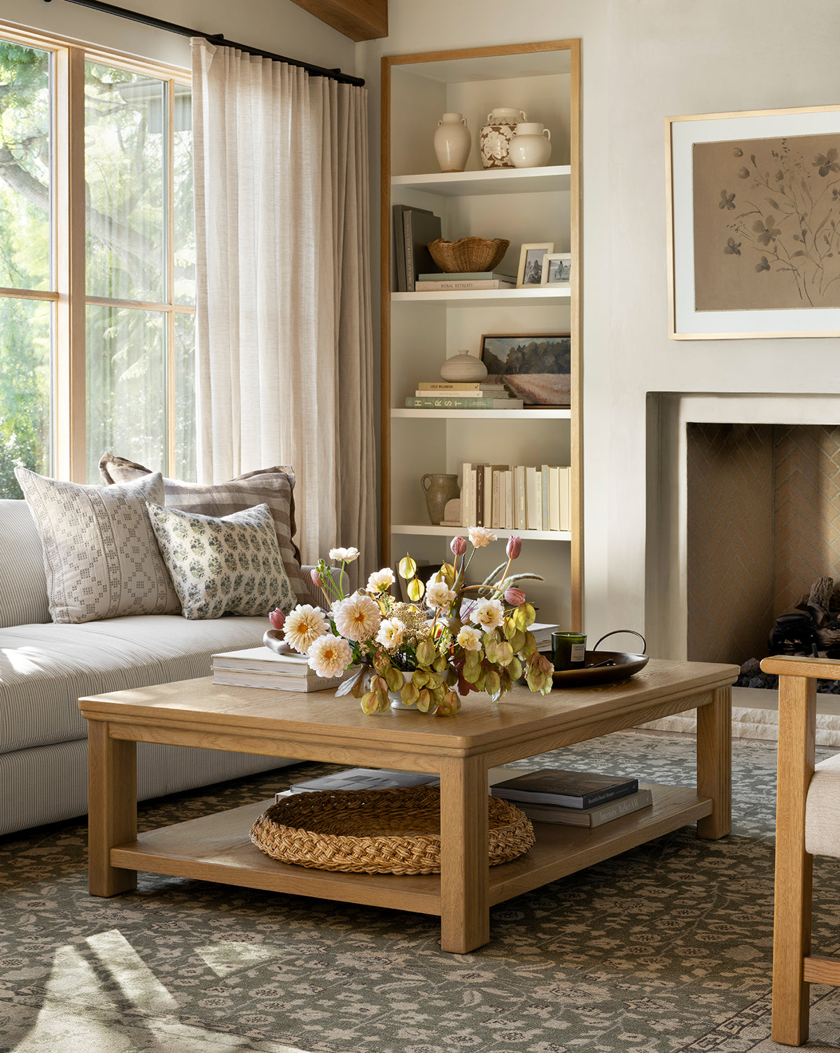 A cozy living room with a light sofa, patterned pillows, an Ambrose Coffee Table by YANG CHENG WOODEN INDUSTRIES INTERNATIONAL (VIETNAM) CO LTD topped with flowers, a woven tray beneath, built-in shelves, fireplace, and large window with sheer curtains. Vendixs