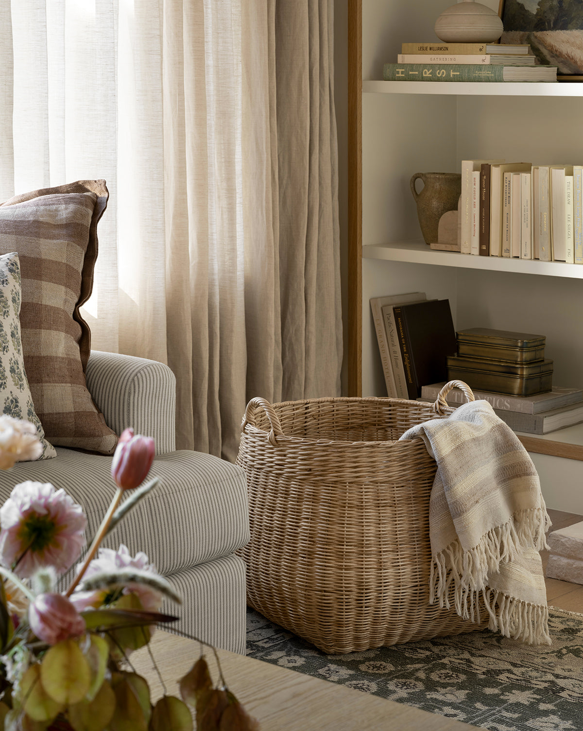 A cozy living room corner with a striped armchair, a plaid pillow, the Boyce Woven Basket holding a fringed blanket, books on white shelves, sheer curtains filtering soft natural light, and pink flowers in the foreground, by Vendixs