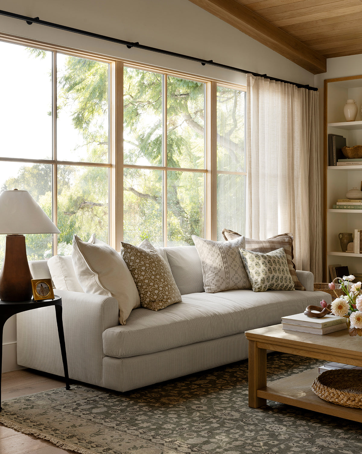 A bright living room features the Makerspalm Winthrop Sofa 90 with patterned pillows, a wooden coffee table with flowers, a side table and lamp, built-in decor shelves, light curtains, and a patterned rug for a cozy finish from Vendixs