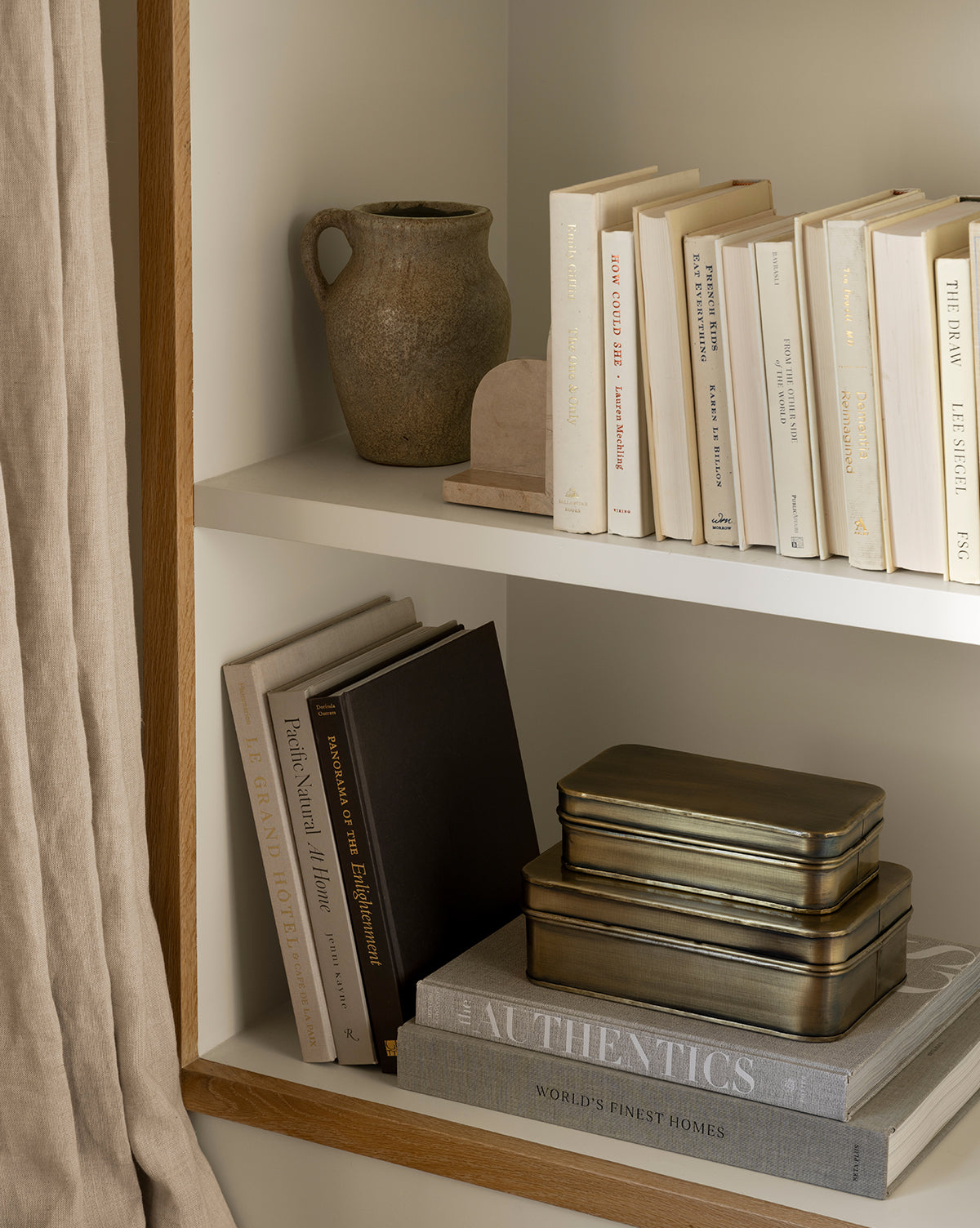 A bookshelf holds neutral-toned books, two stacked metal tins, a small arch decor piece, large hardcover architecture books, and features the Sabine Pitcher Vase. Beige curtains hang to the side, by Vendixs