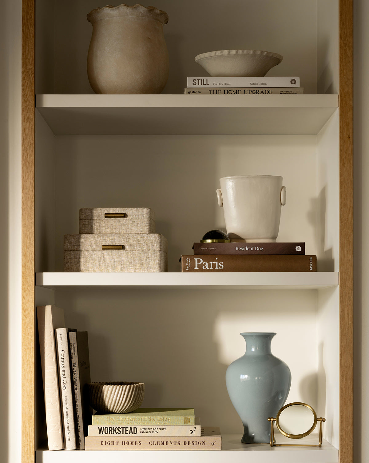 A neatly arranged bookshelf features Swansea Indoor Pot, ceramic vases, stacked books, decorative boxes, and small sculptural objects on three shelves in a warmly lit setting, by Vendixs