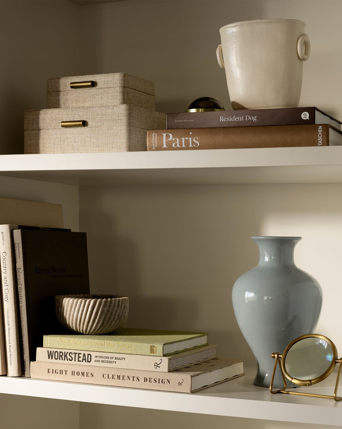 A white bookshelf neatly displays books, beige storage boxes, a cream earthenware vase, the Natalia Vase in light blue, a small bowl, and a gold magnifying glass on two shelves, by Vendixs