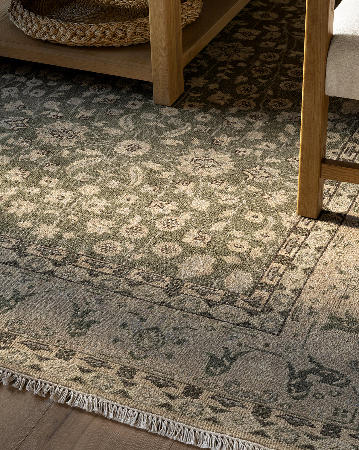 A close-up of the Fernwood Hand-Knotted Wool Rug, showcasing floral patterns in muted green and beige. The rug is accented by a wooden table with a woven basket beneath and a chair, all bathed in natural light, by Vendixs