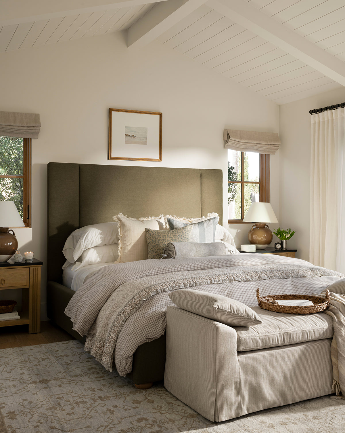 Cozy bedroom featuring the Makerspalm Mina Bed with layered neutral bedding, bedside tables and lamps, beige curtains, soft lighting, a cushioned bench at the foot of the bed, and framed art on the wall by Vendixs