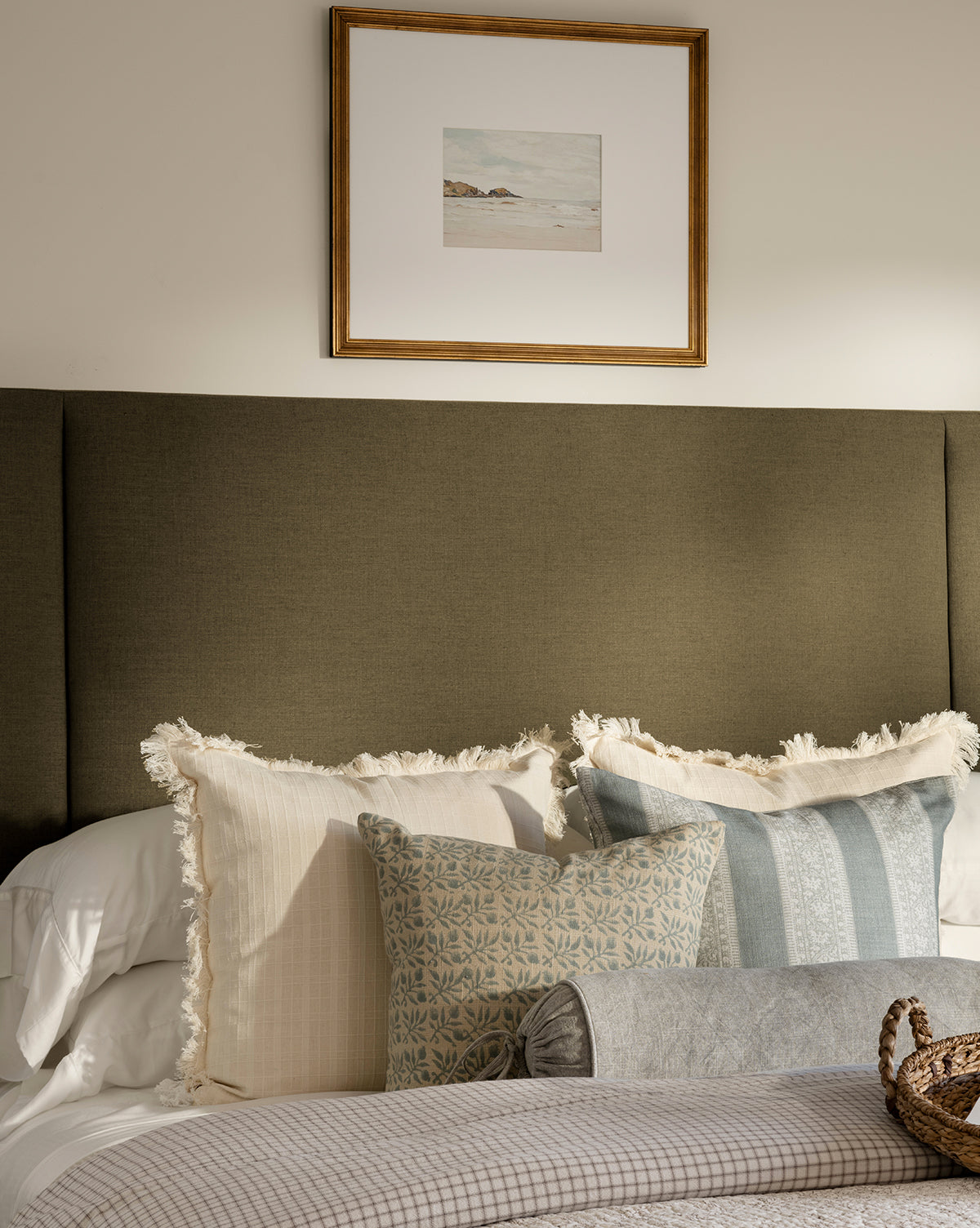 A neatly made bed with layered neutral-toned pillows sits against an olive green upholstered headboard. Above the bed, a framed giclee print titled Shoreline Rocks hangs on a light-colored wall by Vendixs