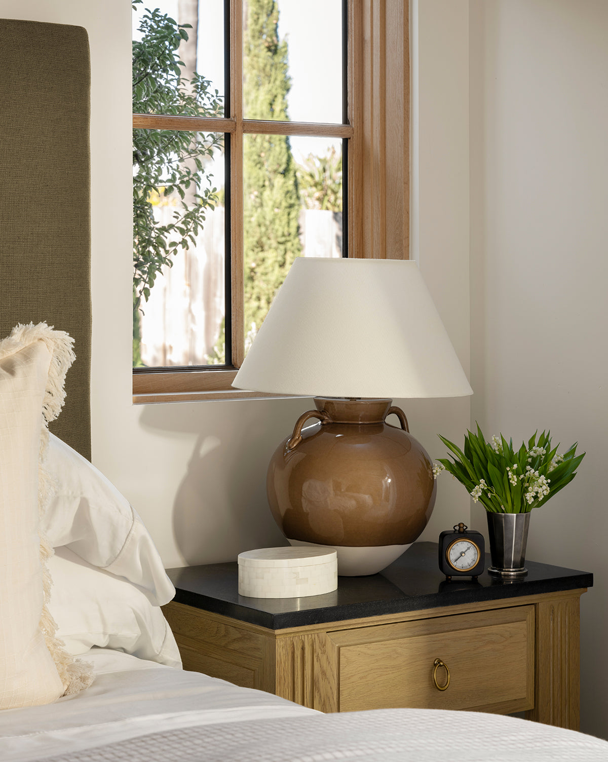 A Derby Vase with elegant flower arrangements joins a brown ceramic lamp, small clock, and round marble box on a bedside table beside white bedding. Sunlight streams through the window behind the table, by Vendixs