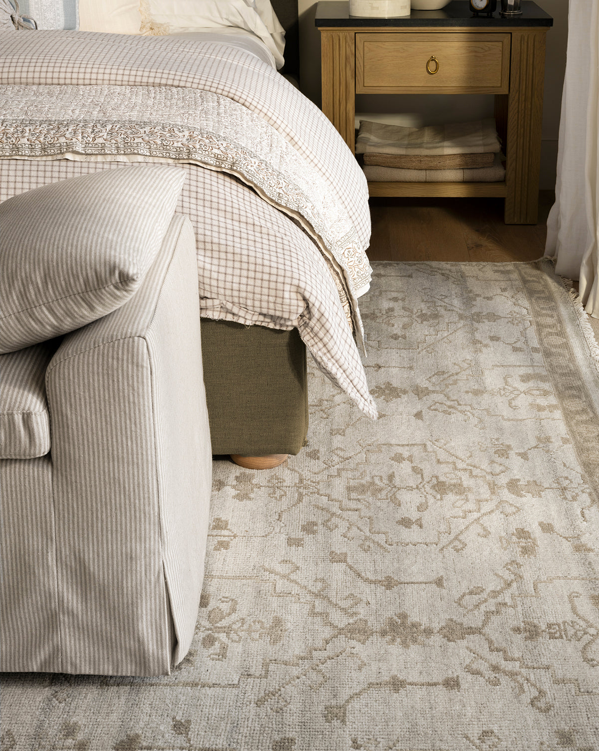 A cozy bedroom corner with a striped armchair, a neatly made bed with plaid and lace bedding, folded blankets on the nightstand shelf, and the Lexington Hand-Knotted Wool Rug crafted by artisans, by Vendixs