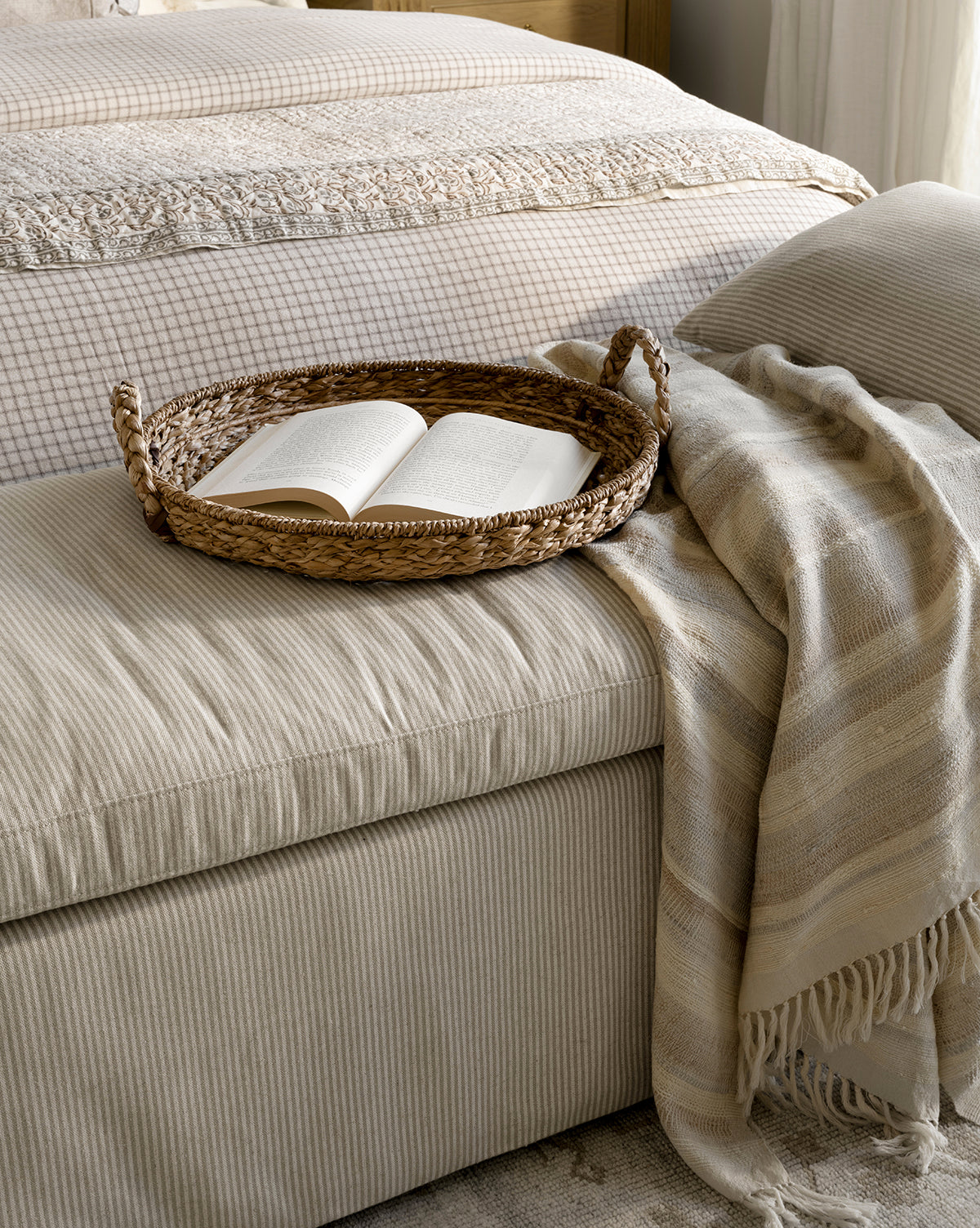 The Salter Woven Tray with an open book sits on a beige, textured ottoman next to a cozy bed. A light, striped throw blanket is draped over the ottoman, adding warmth and inviting comfort, by McGee & Co.