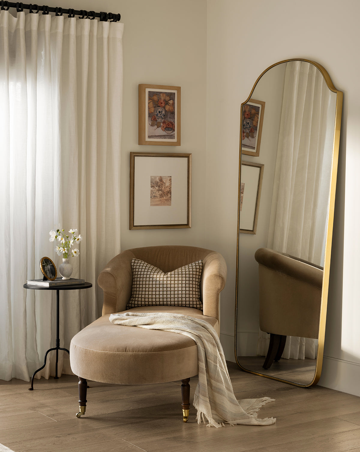 A cozy beige Makerspalm Clemence Chaise with checked pillow and striped throw sits by a round black side table. An arched mirror, framed art, and tall curtains on cream walls create a warm, elegant corner—styled by Vendixs