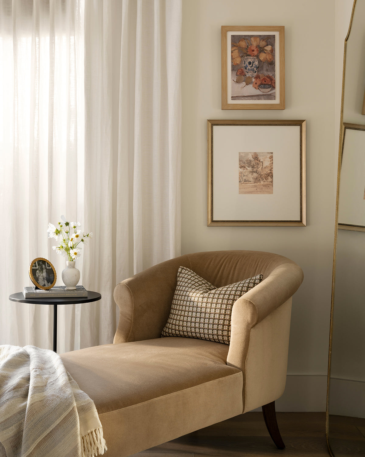 A cozy reading nook features a beige chaise lounge, patterned pillow, striped blanket, round side table with flowers and photo, white curtains, and the Harewood Park print in a distressed silver frame on the cream wall, by Vendixs