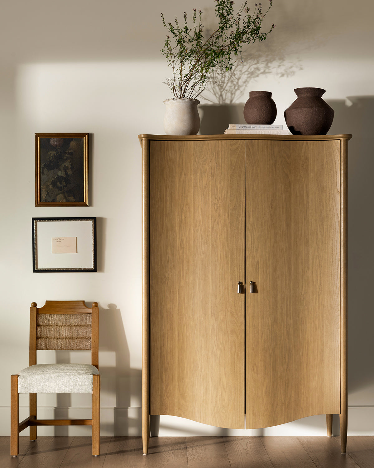 A wooden cabinet with curved edges stands against a beige wall, topped with books, two brown vases, and a plant. Next to it is the Alberta Dining Chair featuring a white cushion. Three framed pictures hang on the wall above, by McGee & Co.