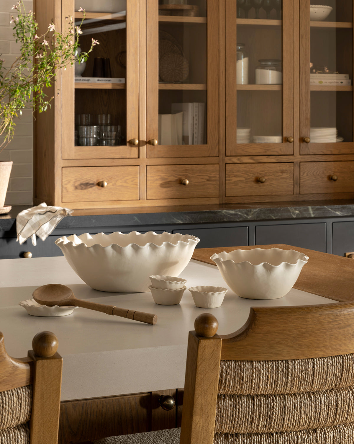 A kitchen with wooden cabinets, glass doors, and a marble countertop features a white island displaying nested Olindo Serving Bowls (Set of 2) with scalloped edges, matching cups, a wooden spoon, and two woven-back chairs by Vendixs