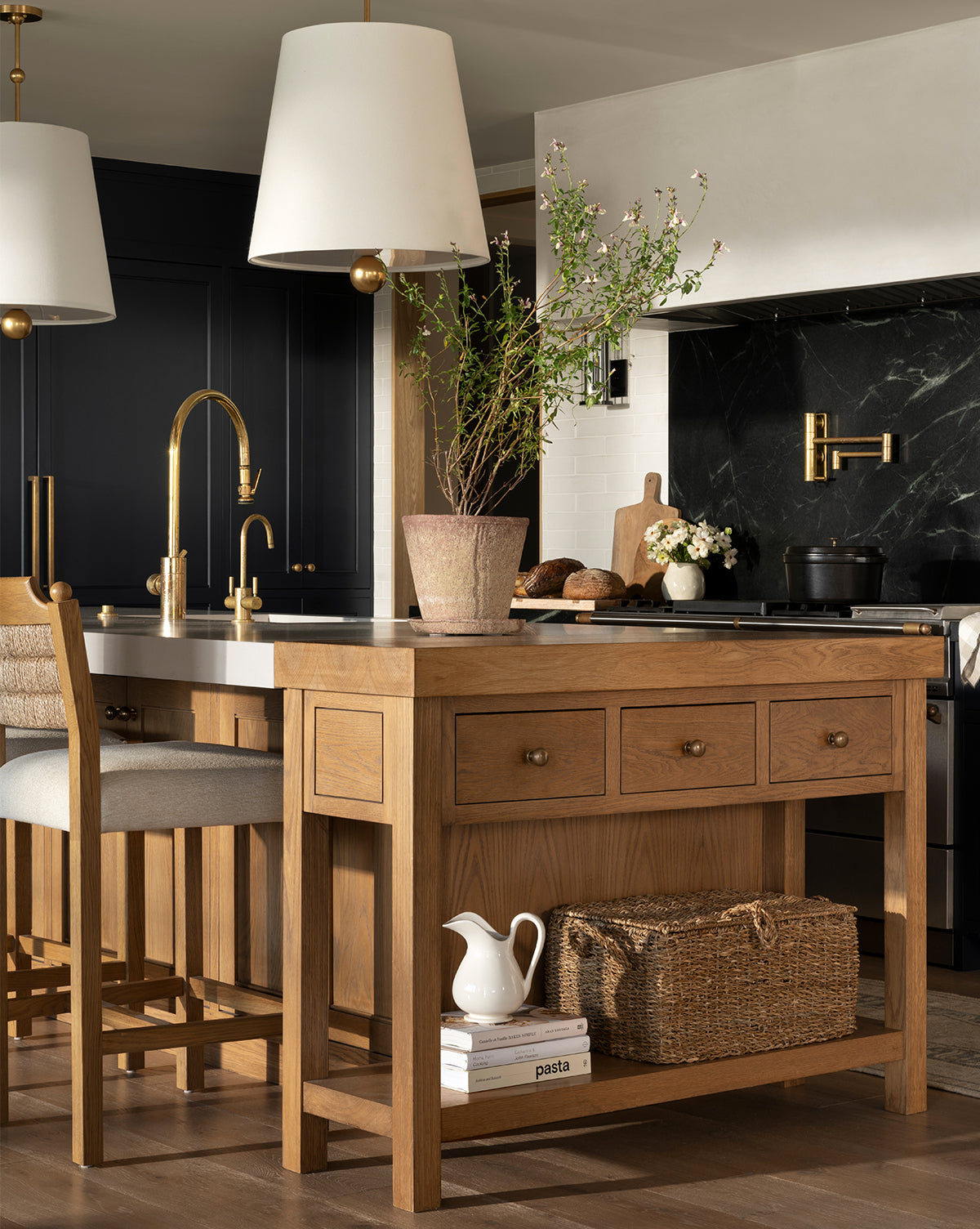 A modern kitchen features a wooden island, gold fixtures, two pendant lights, black cabinetry, a potted plant, wicker basket, and the Lumen Pitcher in white finish on the island shelf near the pasta jar. Marble backsplash and stove in background, by Vendixs