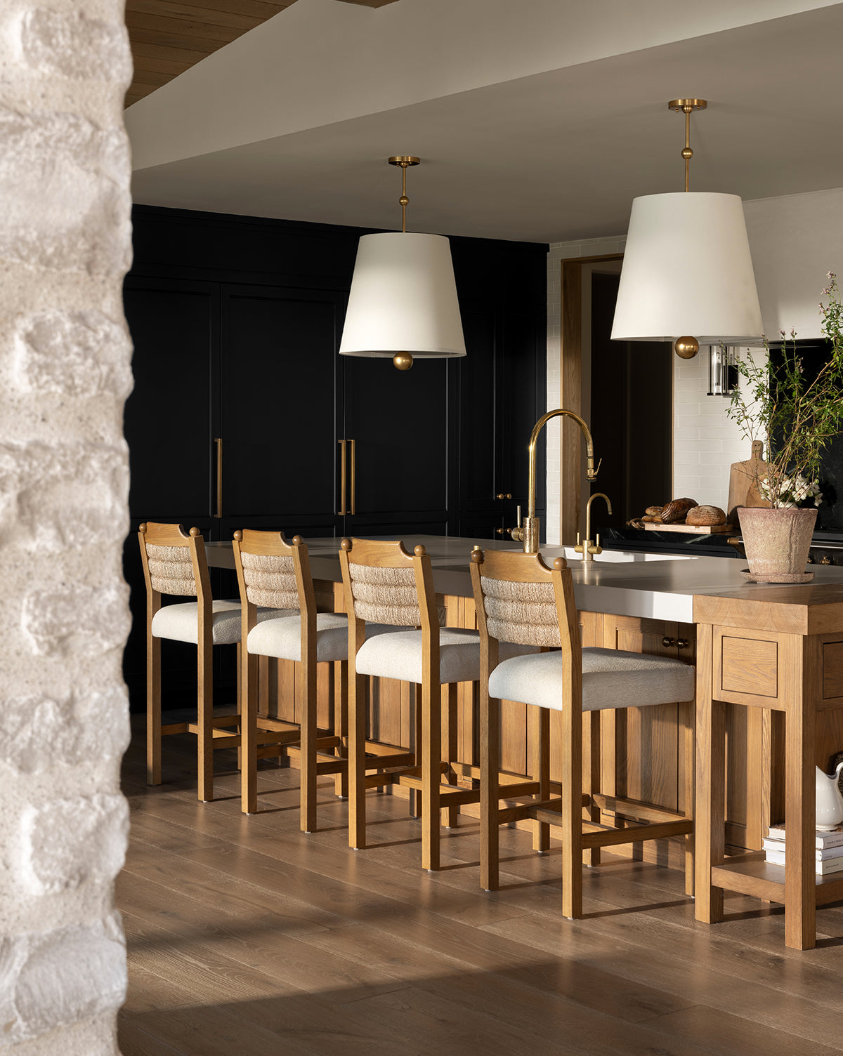 A modern kitchen features a honey oak island, four Makerspalm Alberta Counter Stools with cushions, gold fixtures, black cabinets, two large white pendant lights above the counter, and a potted plant on the island. Vendixs