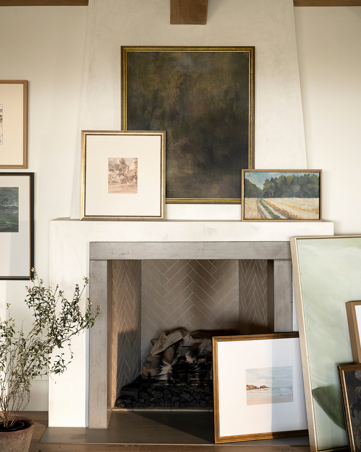 A white fireplace with a herringbone brick interior displays framed artwork, including the Around the Bend forest path print, leaning on the mantel and hearth, by Vendixs A plant to the left complements the simple, elegant decor.