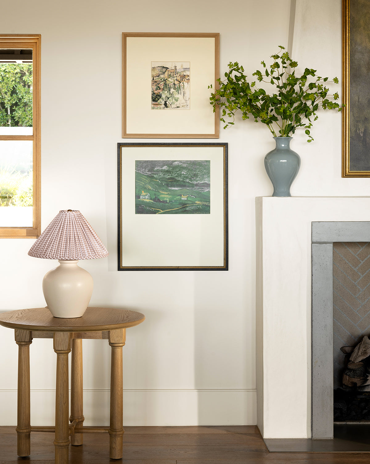 A cozy living space with a light wood side table, a cream ceramic lamp with a patterned shade, framed artwork on the wall, the Natalia Vase with green branches, and a white fireplace beside a window, by Vendixs
