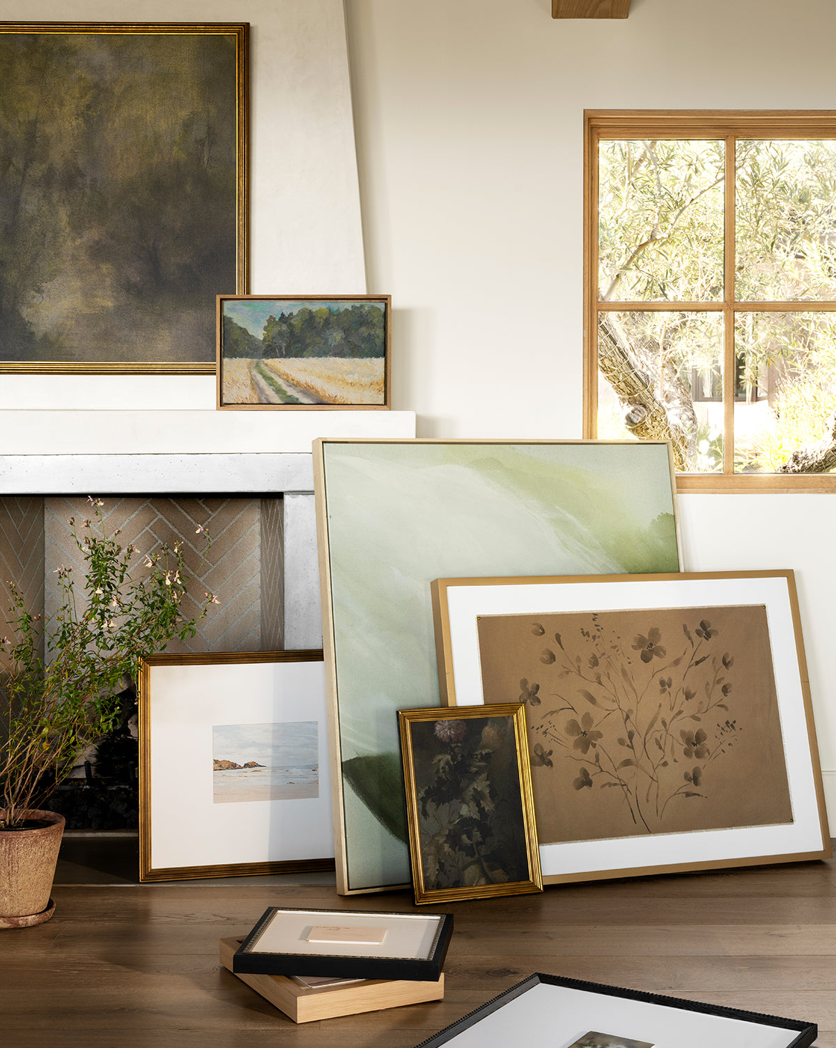 The framed Thistle Down artwork in an antique gold frame leans against a fireplace on the floor of a sunlit room with a window and potted plant, by Vendixs