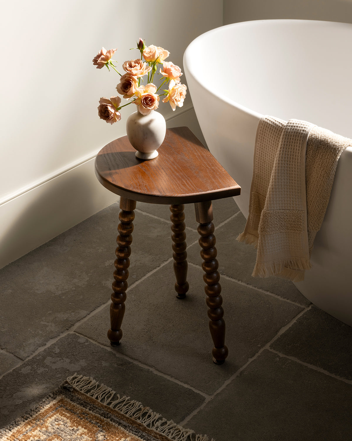 The Alice Wood Stool, featuring three carved ash wood legs, stands beside a white bathtub draped with a beige towel, holding a vase of pink roses on a stone tile floor in a softly lit bathroom, by Vendixs