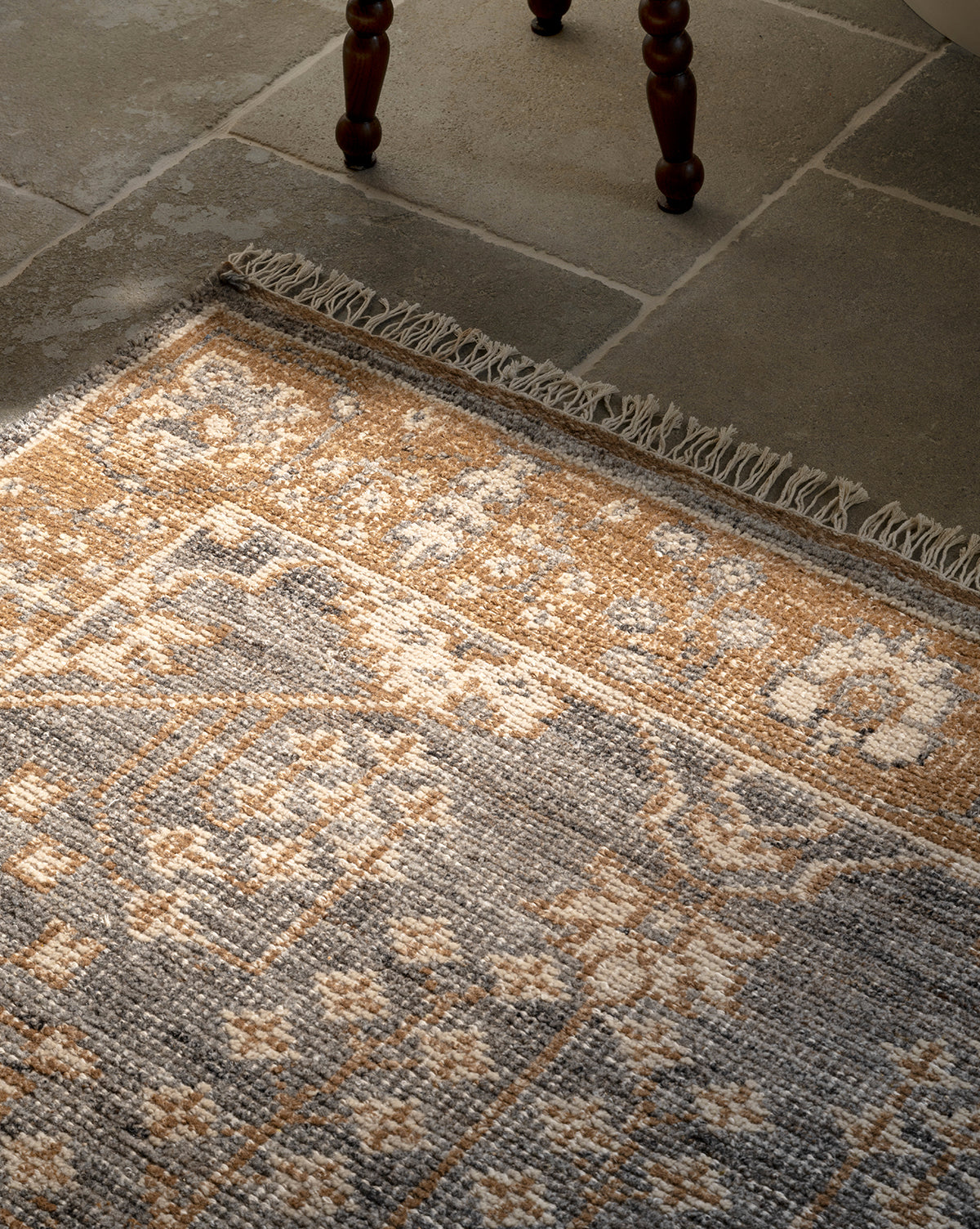 A close-up of the Elison Hand-Knotted Wool Rug in beige, brown, and gray on stone tile flooring, showing artisan details like fringed edges and a wooden chair leg at the top, by Vendixs