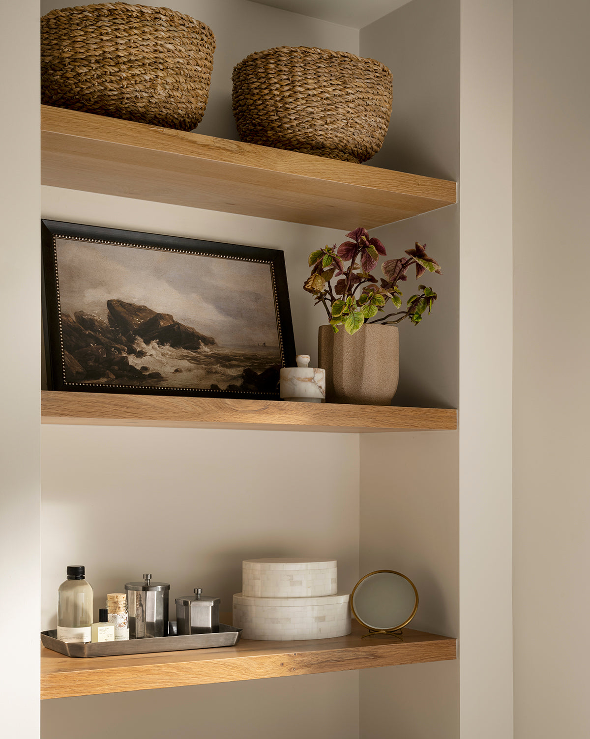 Three wooden shelves display two woven baskets, a Seagrass Footed Bowl, a framed seascape, a beige potted plant, marble jars, toiletries, and a round mirror—highlighting natural accents in this minimalist neutral-toned space.