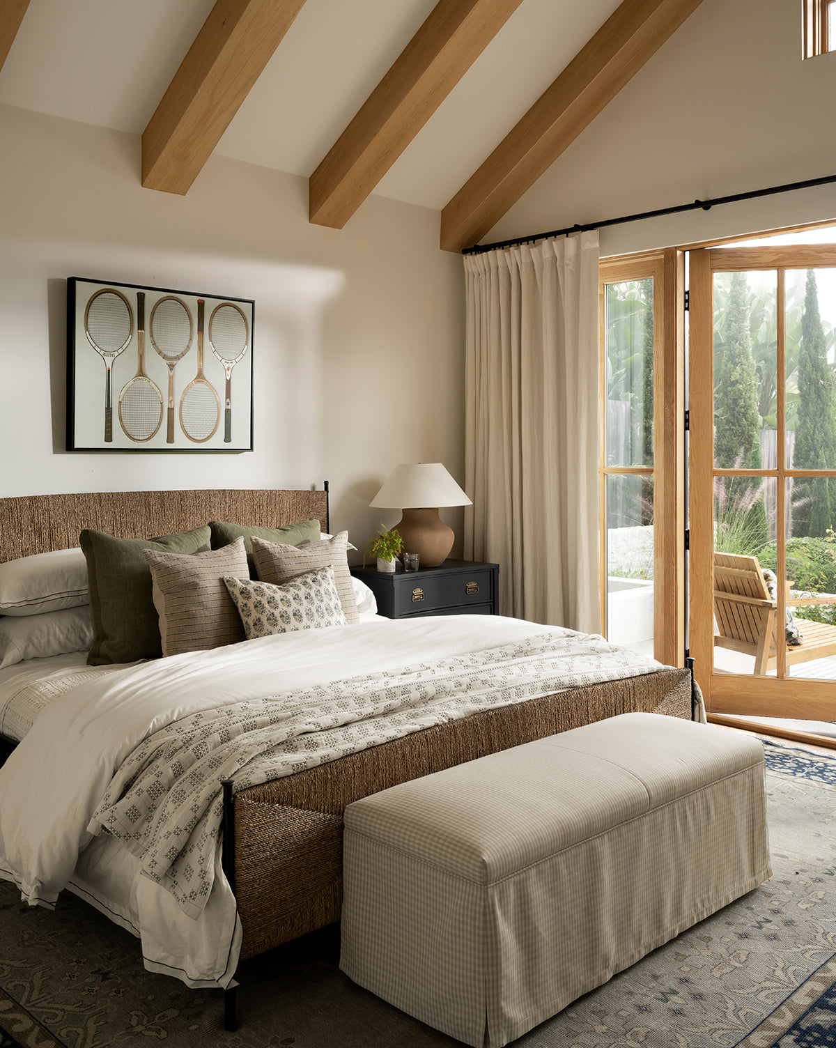 A cozy bedroom with a woven bed frame, neutral bedding, decorative pillows, and a custom Latimer Bench at the foot of the bed. Framed vintage tennis rackets hang above, as large windows fill the space with light and greenery views by Vendixs