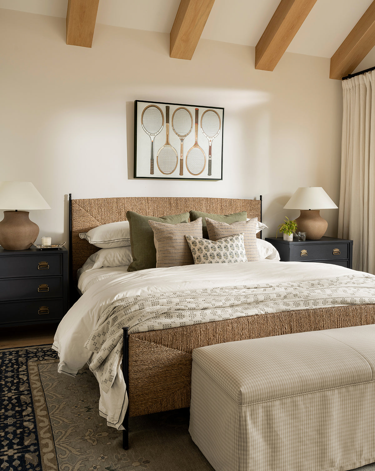 A cozy bedroom featuring the Peregrine Bed with a woven seagrass headboard, layered pillows and blankets, black nightstands with white lamps, exposed wood beams, tennis racket artwork above the bed, and a checkered bench at the foot, by Vendixs