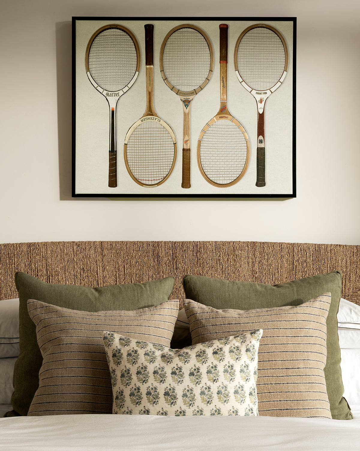 The Rackets 1 giclee print, featuring five vintage wooden tennis rackets, hangs above a neatly made bed with green and striped pillows, a floral cushion, woven headboard, neutral bedding, and a black woodgrain frame, by Vendixs