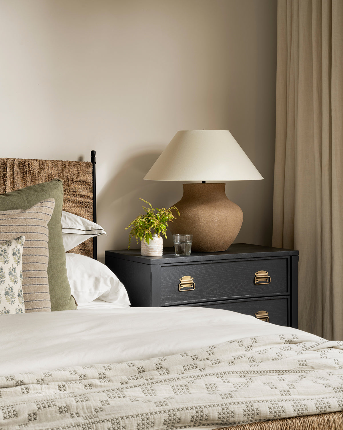 A cozy bedroom corner features a bed with patterned pillows, a black nightstand holding the Parma Table Lamp (textured ceramic base, ivory linen shade), a small plant, and water glasses set against cream walls and beige curtains, by Vendixs