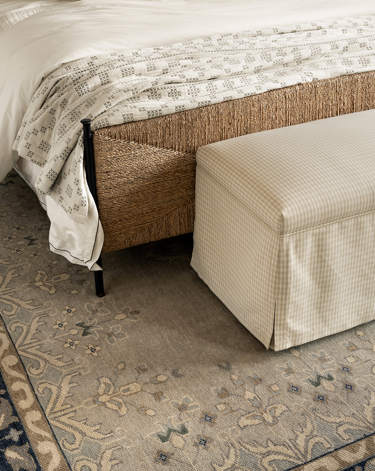 A beige checkered bench sits at the foot of a bed with a woven frame and white patterned bedding, all atop the Deveraux Hand-Knotted Wool Rug in neutral tones, by Vendixs