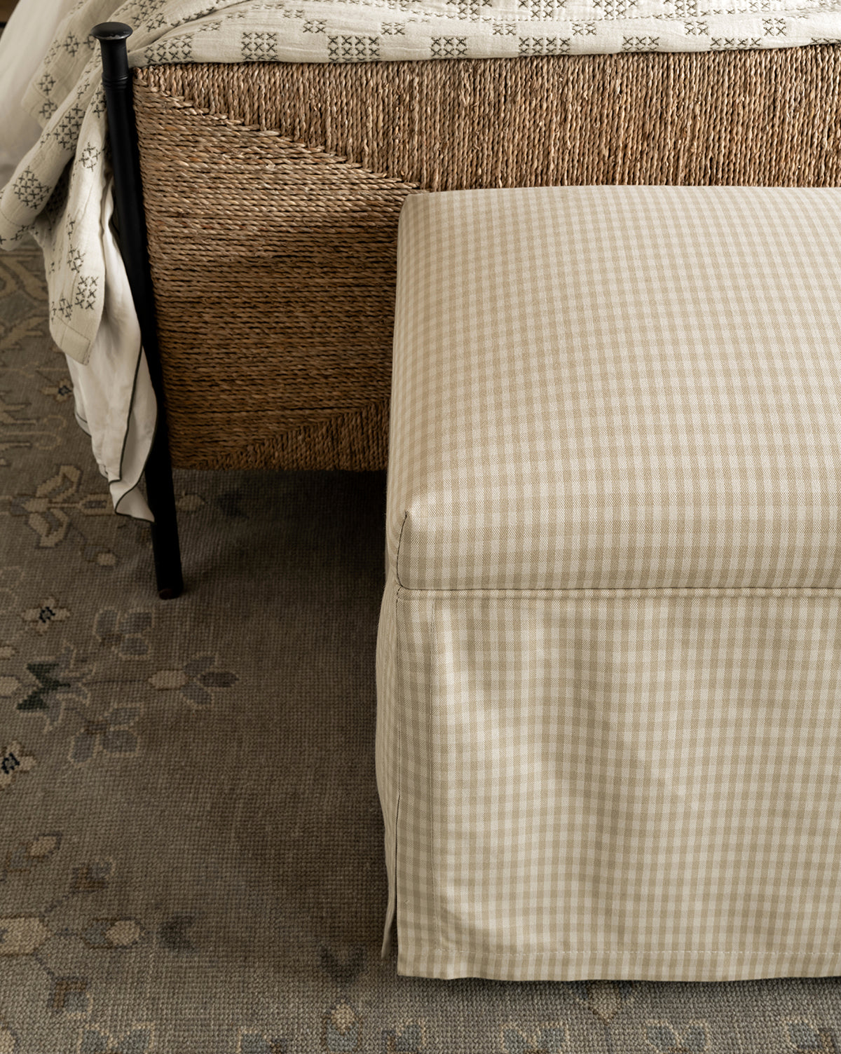 A beige checkered Latimer Bench is placed at the foot of a woven bed with a patterned duvet, atop a floral-patterned rug, by Vendixs